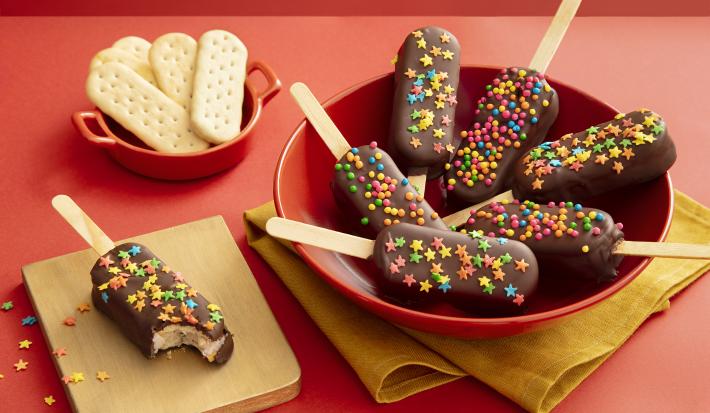 Foto da receita de Pirulito de Biscoito. Observa-se 7 pirulitos decorados com chocolate e confeitos em um prato fundo vermelho sobre um guardanapo de pano amarelo