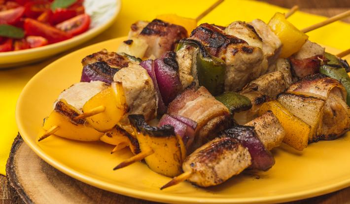 Foto bem aproximada de um prato amarelo sobre o qual há quatro espetinhos montados com carne de porco, cebolas roxa e tradicional e pimentões verdes e amarelos. Ao fundo há um prato branco com uma salada de tomates.