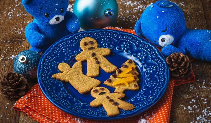 Fotografia em tons de laranja e azul em uma bancada de madeira, um paninho laranja, um prato azul com os biscoitinhos natalinos em cima dele. Ao lado, decoração e enfeites de natal.