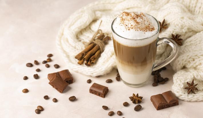 Fotografia em tons de branco de uma xícara de café de vidro com cappuccino feito de café, chocolate em pó, canela e laranja. Por cima do cappuccino tem canela salpicada, ao lado da xícara tem canelas em pau, chocolates, anis estrelado e uma lã branca.
