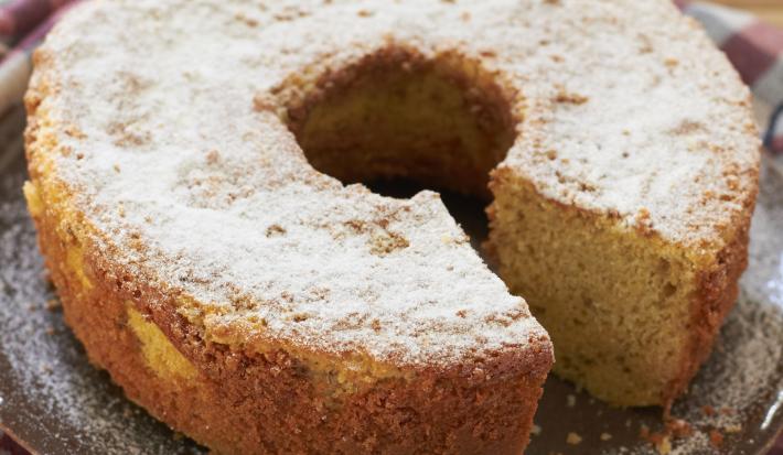 em uma mesa de madeira contém um pano xadrez nos tons vinho e cinza e por cima do pano o bolo de fubá coberto com açúcar de confeiteiro.