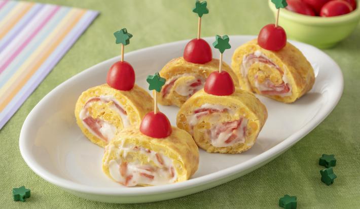 Foto da receita de Enroladinho de Omelete. Observa-se um prato branco com 5 enroladinhos de omelete de pé dispostos. Para prender os enroladinhos, tem um palitinho com estrela verde na ponta espetando uma metade de tomate-cereja.