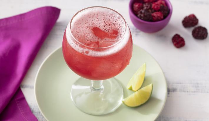 Fotografia em tons de branco, roxo e verde de uma bancada de madeira branca e um prato redondo verde, sobre ele uma taça com o refresco de tônica e duas fatias de limão. Ao lado um paninho roxo e um recipiente redondo roxo com frutas vermelhas.