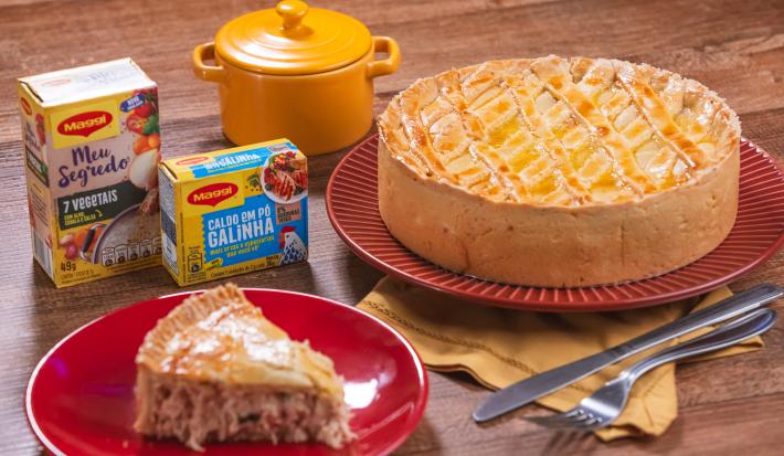 Foto da receita da Melhor Torta de Frango. Observa-se uma torta de frango redonda e grande e, à frente, em um prato de sobremesa, uma fatia dela. Embalagens de MAGGI decoram a foto.