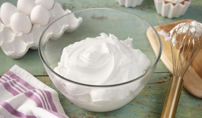 Fotografia em tons de verde em uma bancada de madeira verde, um pano branco com listras roxas, um recipiente de vidro com o merengue dentro dele. Ao fundo, potinhos com docinhos cobertos com merengue, ovos, tábua de madeira com um fouet.