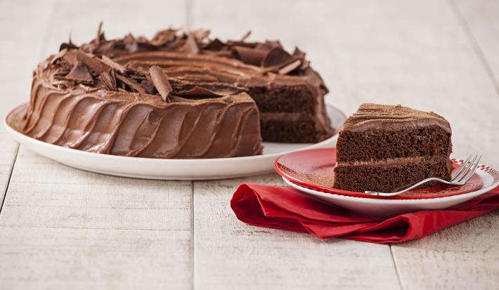 Fotografia em tons chocolate e branco, ao centro prato com bolo de chocolate fatiado, fatia ao lado sobre pratos vermelho e branco com um garfo e sobre guardanapo vermelho, tudo sobre bancada de madeira em pátina.