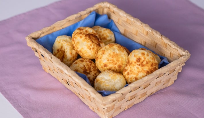 Fotografia de uma cestinha de uma cestinha de madeira retangular sobre um pano quadrado roxo. Dentro da cestinha tem um paninho menor azul com os pães de queijo.