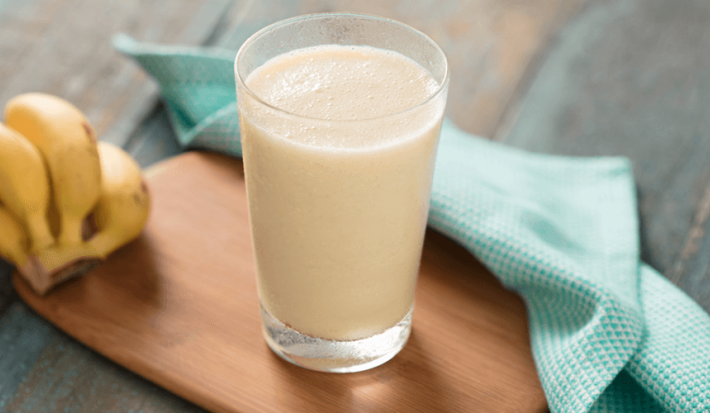 Fotografia em tons de azul e amarelo em uma bancada de madeira escura, uma tábua de madeira, um pano de pano azul com bolinhas, um cacho de bananas e um copo de vidro alto com a vitamina de banana com maracujá e coco.
