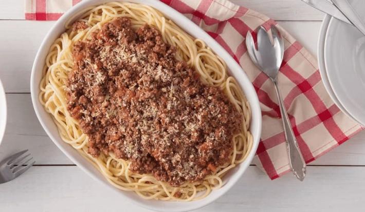 em uma mesa contém um recipiente que comporta o macarrão, ao lado um prato com uma quantidade de macarrão. Ao lado do recipiente contém um pano nas cores vermelho e rosa, e ao lado desse pano dois pratos com dois garfos por cima.