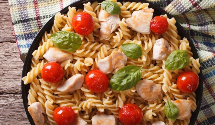 fotografia em tons de bege e vermelho torada de um recipiente redondo preto visto de cima. Contém macarrão tipo parafuso, pedaços de frango e tomates-cereja