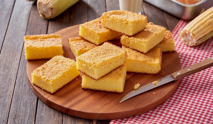 Imagem de uma tábua redonda com pedaços de bolo de fubá cremoso e uma faca em cima. Ao lado, um pano xadrez vermelho e espigas de milho.