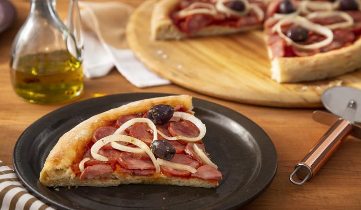 Imagem de um prato preto com uma fatia de pizza de calabresa com cebola com uma tábua de madeira e a pizza ao fundo.