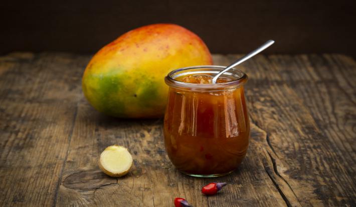 Fotografia de um chutney de manga e laranja temperado dentro de um recipiente de vidro pequeno, como se fosse um copo. Apoiado no chutney, uma colher de chá, e ao fundo, uma manga inteira com casca, sobre uma mesa de madeira.