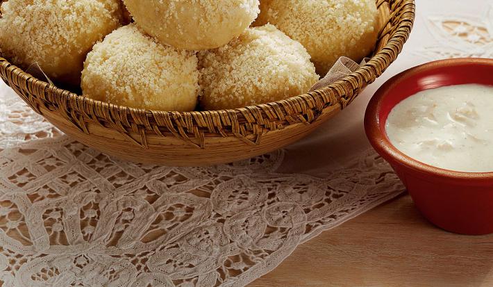 Fotografia em tons de branco, marrom, pardo e vermelho de uma bancada vista de cima. Um pano de renda branco e por cima um recipiente cor pardo e redondo com pães bolinha, ao lado um potinho vermelho redondo com creme branco.