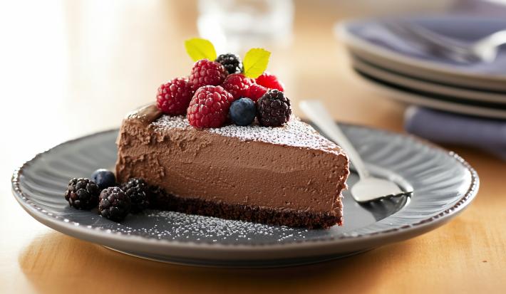 A foto mostra uma fatia da tortinha com uma base de biscoito de chocolate, recheada com creme de avelã e chocolate meio amargo. O topo da torta é coberto por frutas vermelhas frescas, como morangos, amoras e framboesas.