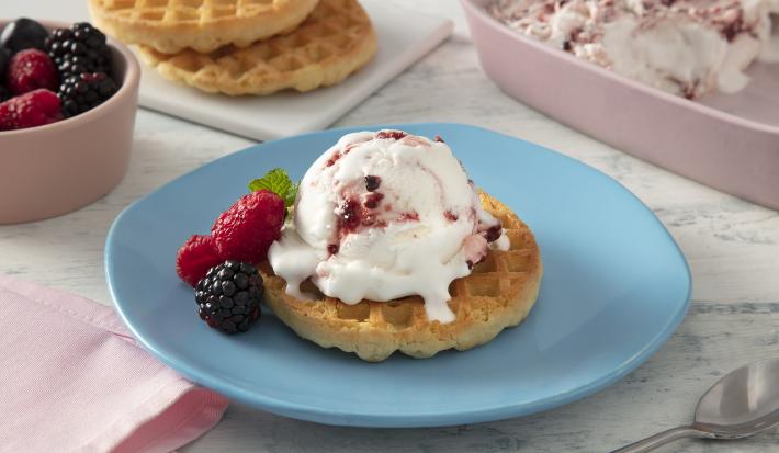 Foto da receita de MOÇA Sorvete de Ninho com Frutas Vermelhas. Observa-se uma bola grande de sorvete sobre um waffle, em um pratinho azul. Frutas vermelhas decoram a receita!