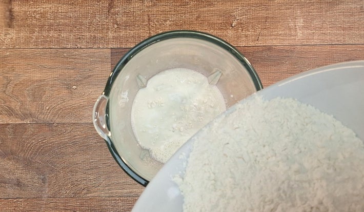 Ingredientes no liquidificador. Uma mão derruba farinha de uma tigela grande dentro do liquidificador.