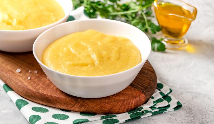 Na fotografia contém um pano branco com bolinhas verdes, por cima uma tábua de madeira de ponta redonda. Acima da tábua dois potes brancos com polenta dentro e ao lado um potinho com azeite.