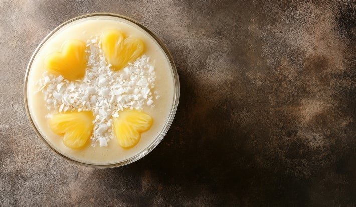 Fotografia vista de cima de um recipiente pequeno, tipo copo de vidro pequeno, com a sobremesa da receita, pedaços pequenos de abacaxi em formato de coração e um pouco de coco ralado. O recipiente está sobre uma mesa escura.