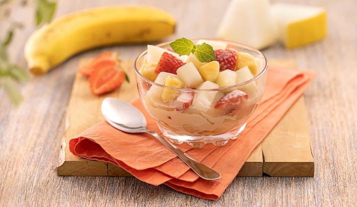 Foto da receita de salada de frutas servida em uma taça de vidro decorada com uma folha de hortelã, sobre um pano laranja com uma colher prateada ao lado. Ao fundo, há morangos, uma banana e fatia de melão