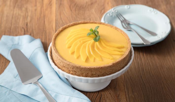 fotografia em tons de marrom, branco e amarelo de uma bancada marrom vista de frente, contém uma bailarina para servir sobremesa branca com uma torta por cima. Ao lado esquerdo contém um prato redondo branco e do lado direito um pano azul com uma espátula