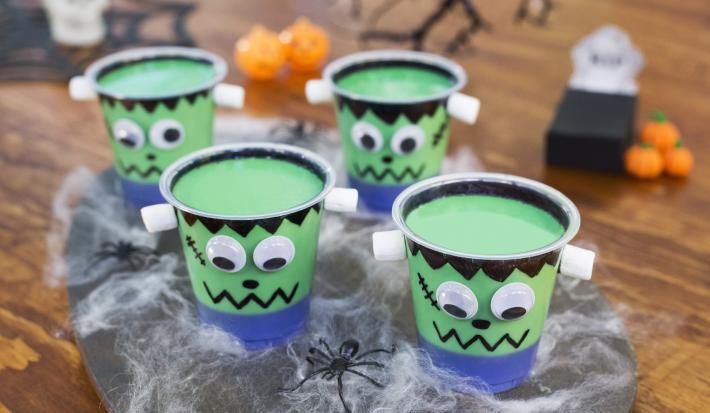 Imagem aproximada de três copos com a receita em tom verde, tudo decorado com tema de dia das bruxas, sobre uma bancada de madeira também decorada com teias falsas, aranhas e outros itens