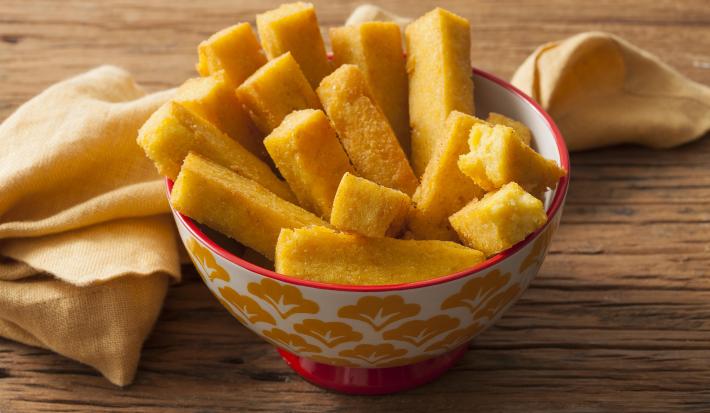 Fotografia em tons de amarelo de uma bancada de madeira com um pano amarelo retorcido e um potinho decorado branco, amarelo e vermelho, com vários palitos de polenta frita dentro.