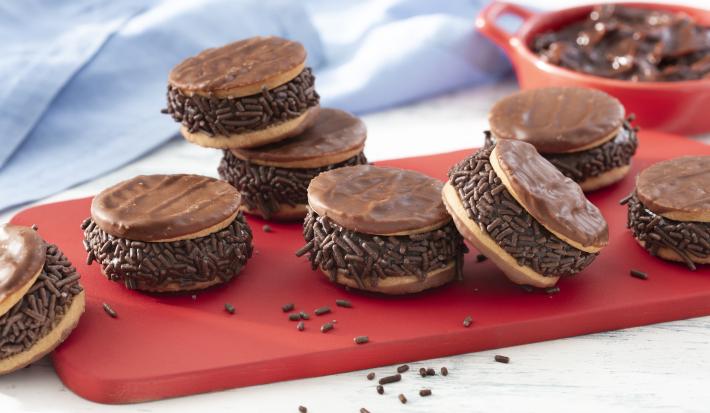 Fotografia em tons de vermelho em uma bancada de madeira clara, um pano azul, uma tábua vermelha com vários sanduíches de biscoito calipso recheados com brigadeiro de pão de mel. Ao fundo, um potinho vermelho com o brigadeiro dentro.