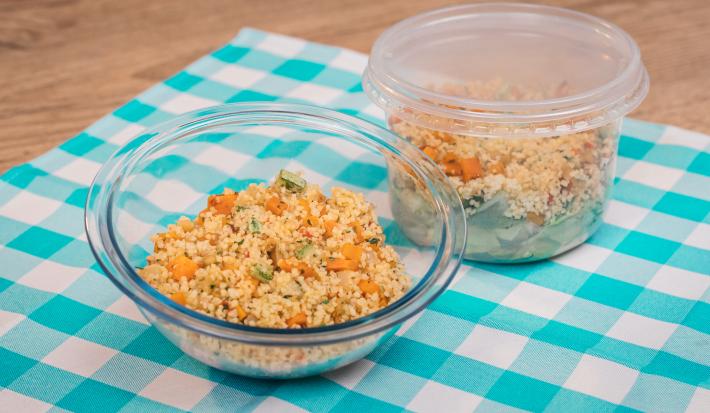 Foto de um pote de vidro redondo e um pote de plástico, ambos com a receita de salada de couscuz marroquino. Os dois estão sobre uma pano azul e branco quadriculado em uma bancada de madeira