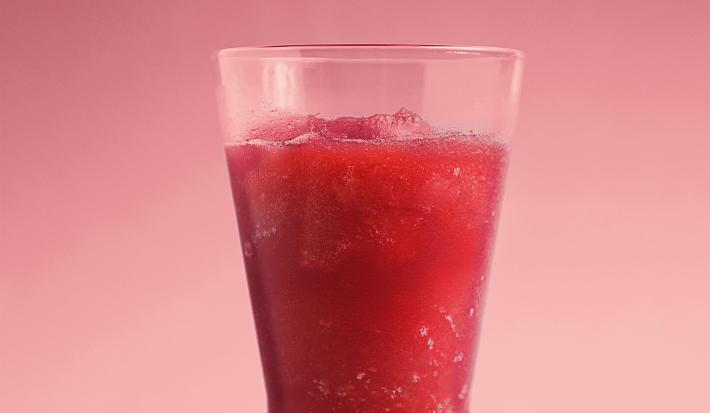 Fotografia em tons de vermelho e rosa de uma bancada rosa sobre ela um copo de vidro com suco de melancia.