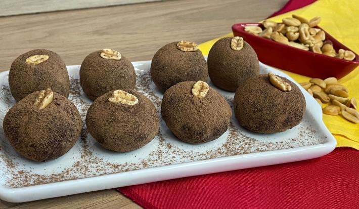 Fotografia em tons de amarelo e vermelho com um prato branco retangular ao centro em uma bancada de madeira. Dentro do prato existem 8 trufas de chocolates enfeitadas com amendoim. Ao fundo existe um pequeno pote cheio de amendoim sem casca.