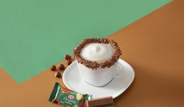 Foto da receita de cappuccino kit kat servida em uma xícara branca com um kit kat avelã ao lado em fundo marrom e verde