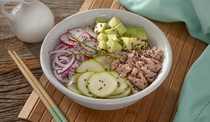 Foto em tons coloridos da receita de poke funcional de atum servida em um bowl de porcelana branco sobre uma esteira de madeira com dois hashis ao lado. Ao fundo há um pano azul claro