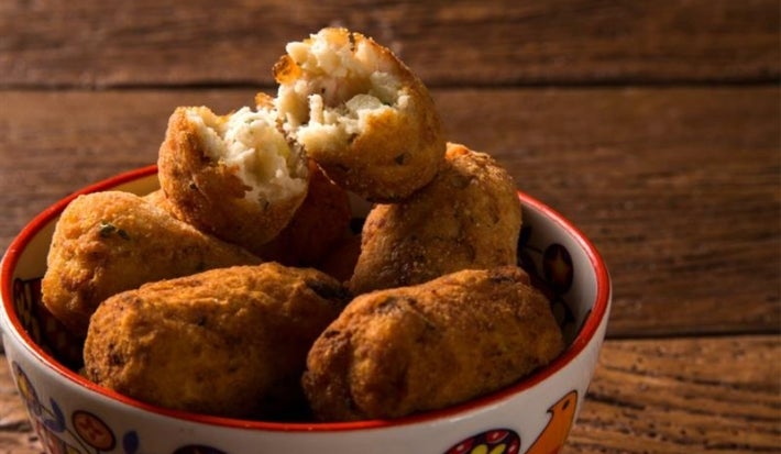 Foto aproximada da receita de Bolinho de Pirarucu, servido em um pote sobre uma bancada de madeira. O Bolinho de cima está aberto pela metade mostrando o interior