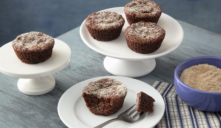 Foto em tons de cinza da receita de bolo de chocolate de farinha láctea aveia servida em diversas porções, três em uma base de porcelana branca e uma com uma garfada servida em um prato de mesmo material. Tudo isso sobre uma mesa cinza