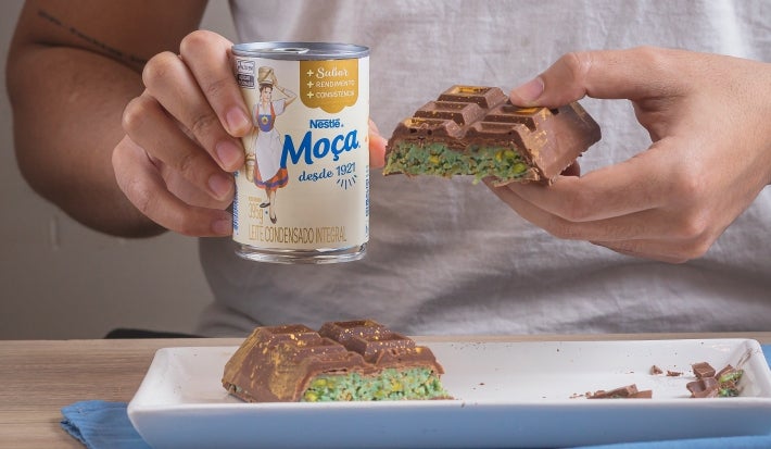 Barra de Dubai, receita de uma barra de chocolate recheada com um creme de pistache e macarrão, decorada com pó dourado e servida em um prato branco com uma pessoa ao fundo segurando metade da receita e a lata de Leite Moça na outra mão