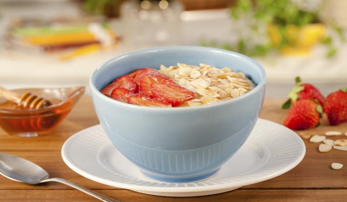 Imagem de um bowl azul claro com mingau de aveia, rodelas de morango, lascas de amêndoas e mel. Ao redor temos morangos, uma colher e um potinho de mel.