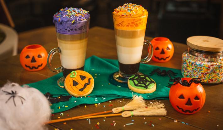 Fotografia em tons de laranja de uma mesa de madeira, duas bebidas, uma decorada com chantilly roxo e uma decorada com chantilly laranja. Em volta biscoitos decorados de abóbora, teia de aranha e elementos de halloween.