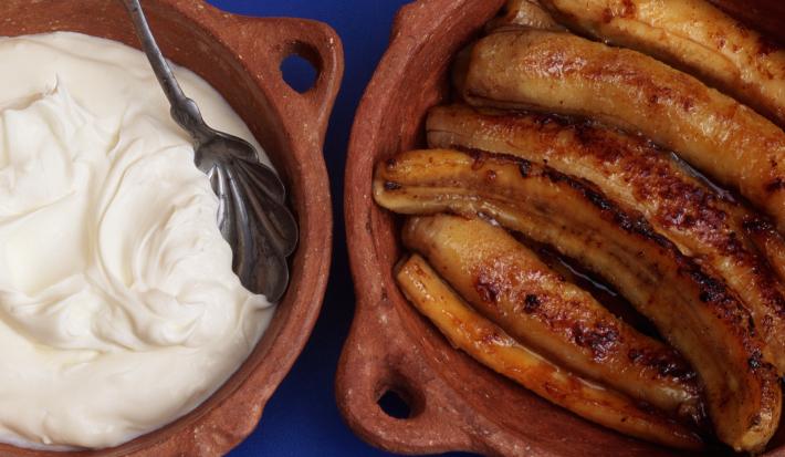 Fotografia vista de cima de bananas cortadas ao meio flambadas dentro de um recipiente marrom de cerâmica. Ao lado esquerdo, um creme branco com consistência mais densa dentro do mesmo recipiente, e dentro do creme, uma colher de prata apoiada.
