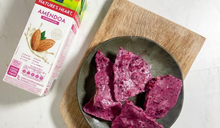 Foto em tons de roxo da receita de barrinha proteica servida em algumas porções em um prato cinza escuro sobre uma tábua de madeira com uma embalagem de bebida vegetal de amêndoas nature's heart ao lado
