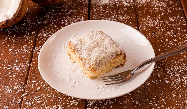 Imagem de um prato branco com um pedaço do bolo coberto com coco e um garfo ao lado.