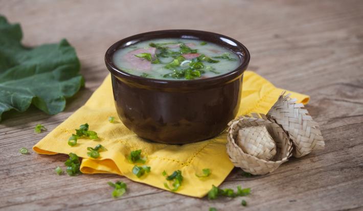 Imagem de um bowl marrom com a receita de caldo verde polvilhada com cebolinha sobre um pano amarelo e chapéus de festa junina ao lado.