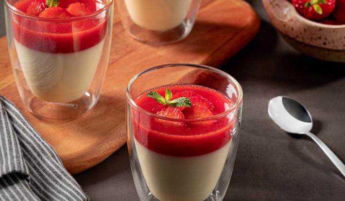 Imagem de três copos pequenos contendo a receita de Panacota com calda de morango. Dois estão sobre uma tábua com um tecido ao lado e um está mais à frente. Ao fundo há um pote com morangos e uma colher de metal