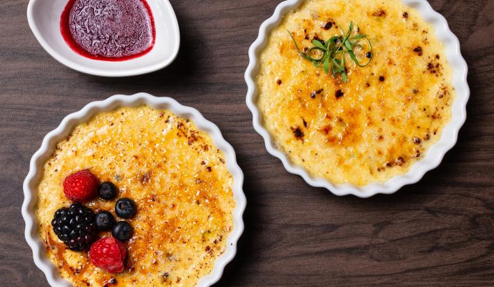 Fotografia vista de cima de dois cremes brulée, um de alho poró com queijo canastra e o outro, sabor cumaru com pistache e frutas vermelhas, os dois cremes brulée estão em ramekins. Ao lado tem uma calda de frutas vermelhas.