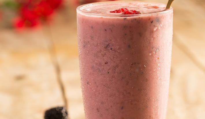 Fotografia em tons de rosa em uma mesa de madeira com um copo de vidro ao centro com o shake de frutas vermelhas dentro. Morangos e mirtilos espalhados pela mesa.