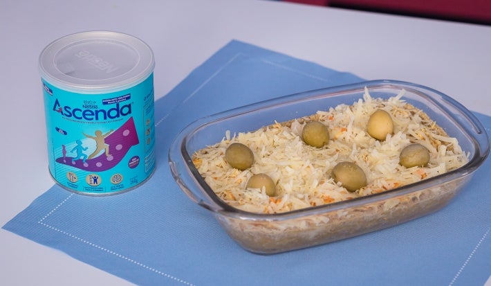 Fotografia de um recipiente refratário de vidro, retangular, ao lado da lata de Ascenda, sobre um paninho azul quadrado. Dentro do recipiente tem a torta com massa de grão de bico e ascenda, finalizada com queijo parmesão e azeitonas.