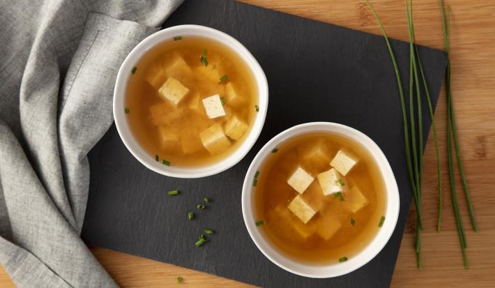 Imagem vista de cima de 2 tigelas brancas com sopa de missoshiro e todu sobre uma tábua preta e pedaços de cebolinha ao lado