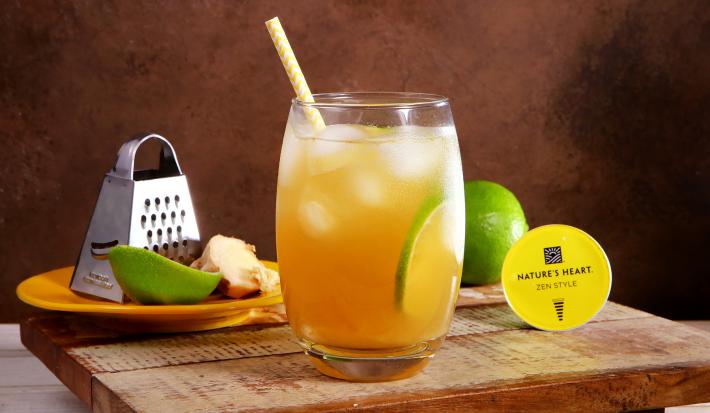 Fotografia em tons de amarelo e verde em uma bancada com uma tábua de madeira clara, um copo de vidro com o chá de gengibre com limão dentro dele. Ao fundo, um ralado, uma fatia de gengibre e um pedaço de limão e cápsula do chá Zen Style Nature´s Heart.