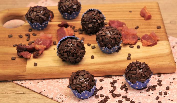 Fotografia em tons de preto em uma bancada de madeira de cor marrom. Ao centro, uma tábua de madeira contendo os brigadeiros em cima. Ao redor, há alguns pedaços de bacon espalhados.