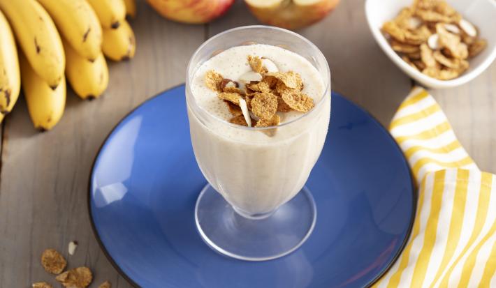 Imagem de um copo com a bebida cremosa em tom claro, decorado com cereal. Ele está sobre um prato azul e, ao redor, há cereais espalhados na mesa, uma penca de bananas, duas maçãs e um tecido listrado amarelo e branco.
